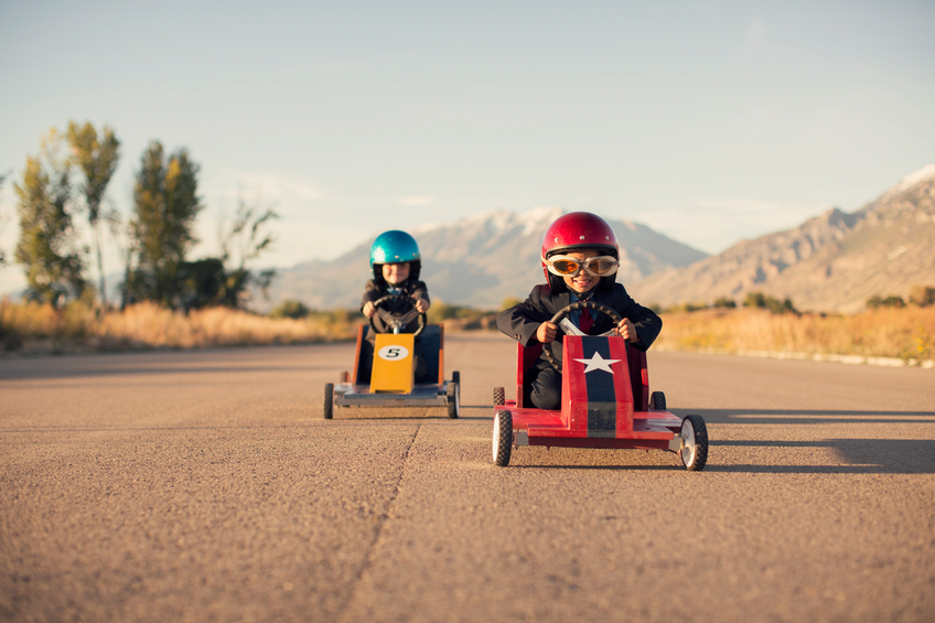 A young boy dressed as a businessman raises his arm in success as his homemade box car is in first place. Both boys are wearing helmets and goggles.